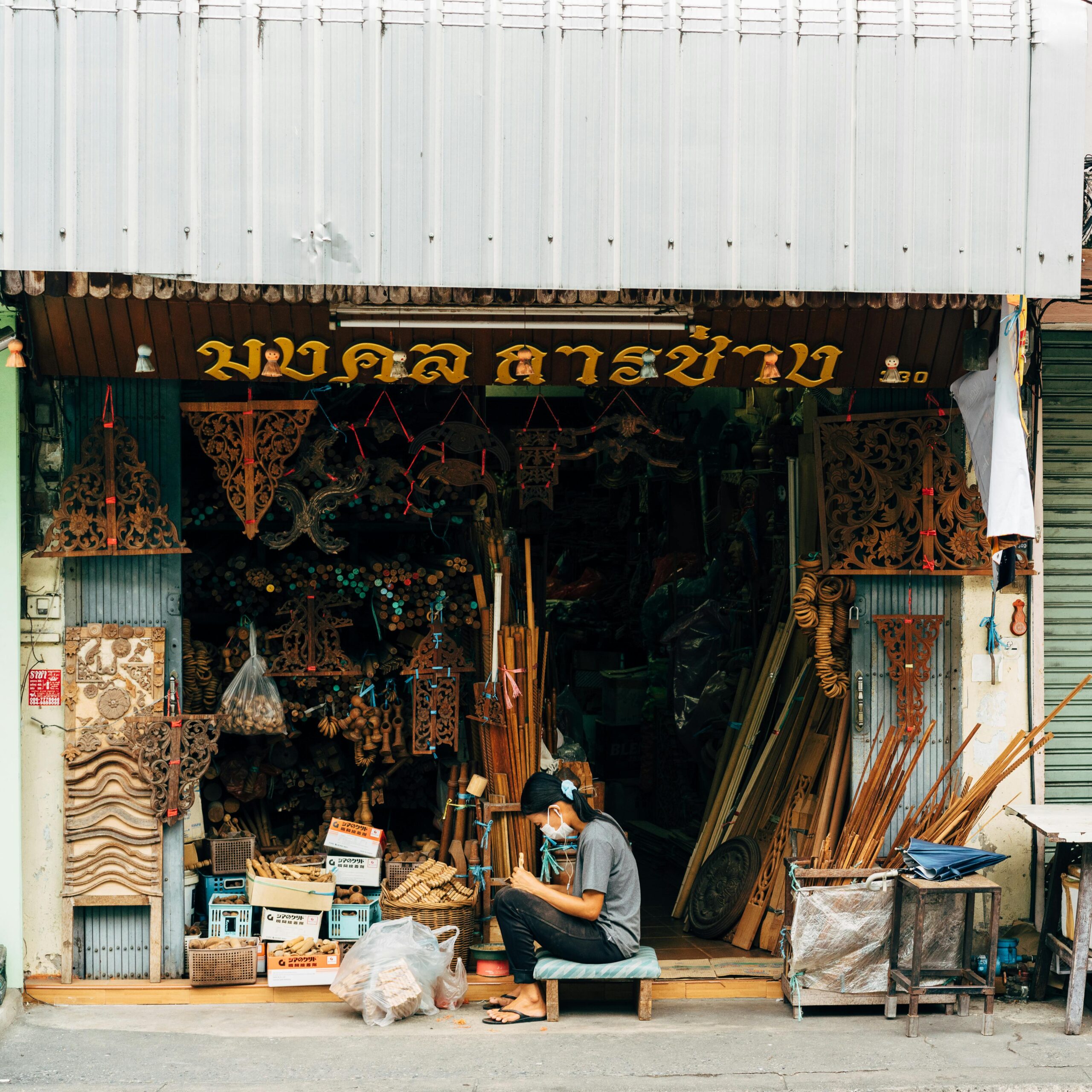 An artisan sits in front of a woodcraft shop in Bangkok, displaying handmade wooden items.